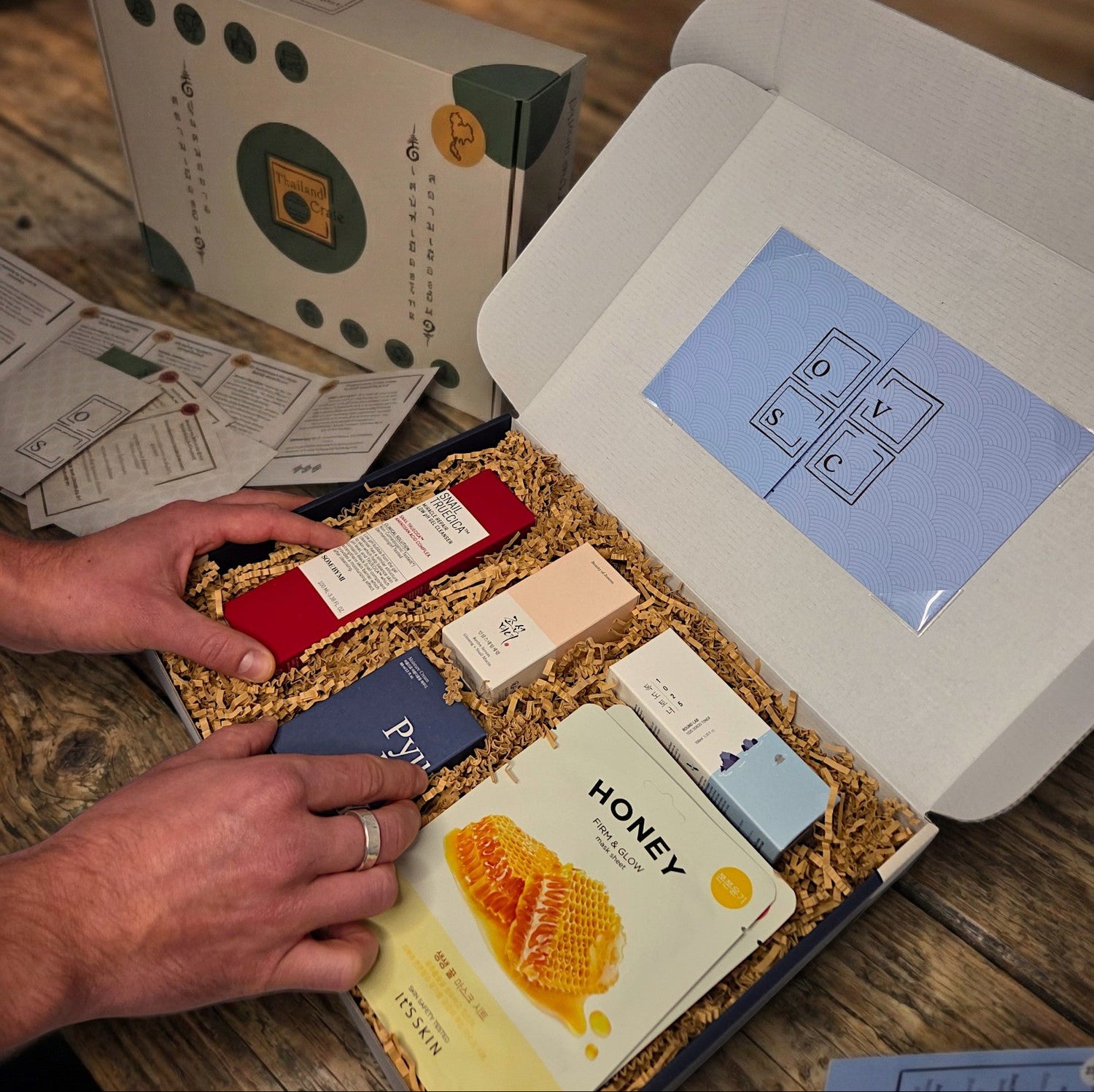 Gestapelte Thailand und Korea Erlebnisboxen von Oversea Crate auf einem rustikalen Holztisch in einem gemütlich eingerichteten Wohnzimmer – die ideale Geschenkidee für eine authentische Auszeit zuhause.