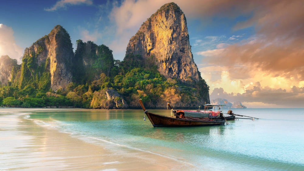 Ein traditionelles thailändisches Longtail-Boot an einem weißen Sandstrand vor imposanten Kalksteinfelsen bei Sonnenuntergang – dein Ticket für eine exotische Auszeit mit der Thailand Erlebnisbox von Oversea Crate.